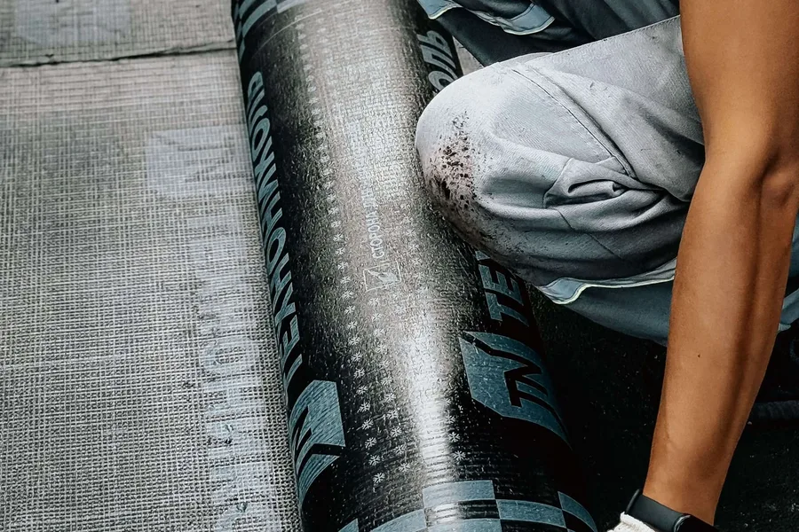 Worker rolling a dark sheet of roofing membrane into position